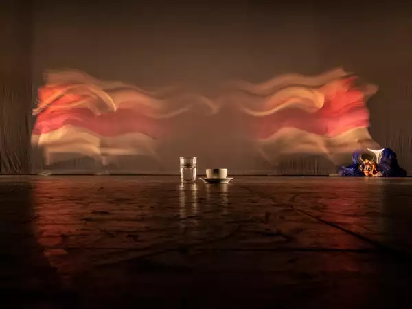 A glass of water and cup and saucer are placed on the floor. Behind them is a curtain and to the right is a puppet.