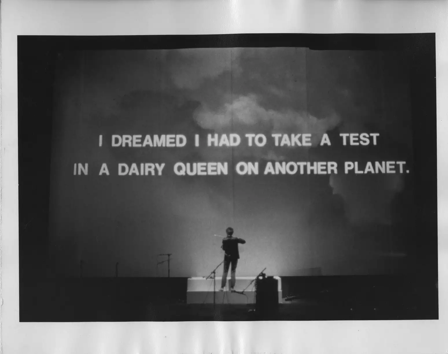 Black and white still from Laurie Anderson's United States Parts 1–4, showing Laurie standing in front of a large screen displaying the words, "I dreamed I had to take a test in a Dairy Queen on another planet."