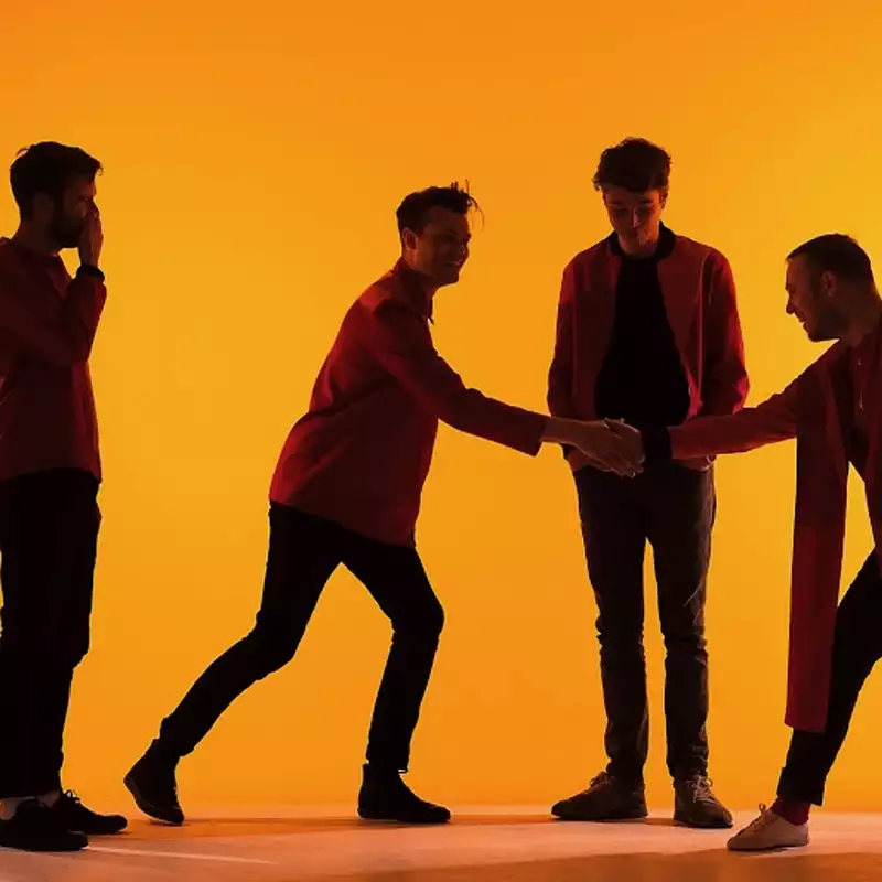 Four members of Everything Everything stand against a vibrant orange background, dressed in red and black, with two engaging in a handshake while the others observe. Their silhouettes and dynamic poses create a visually striking composition.