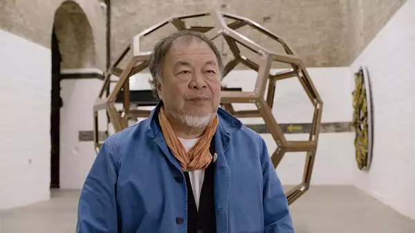 Ai Weiwei talking to the camera in his studio. Behind him is a large circular sculpture.