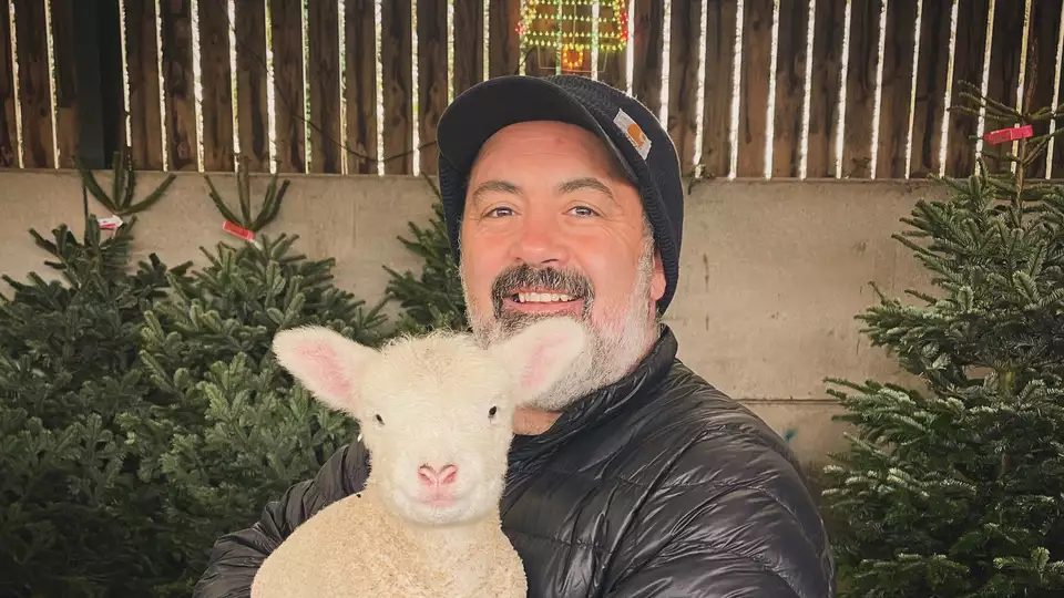 Andy Votel holding a sheep with Christmas trees behind him