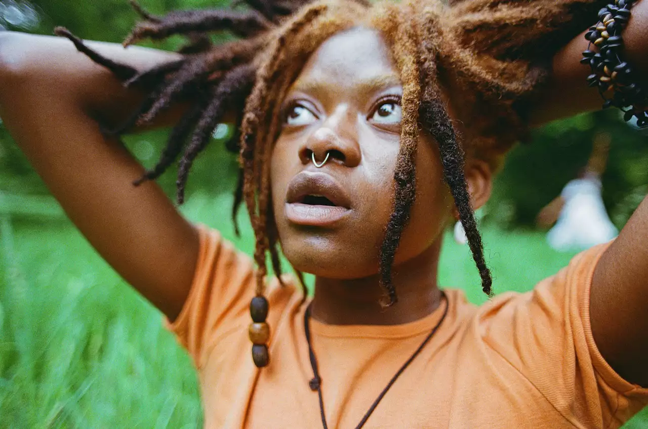 A person in an orange t-shirt with dreadlocks and a nose ring looks upwards, with their hands placed behind their head. There is grass in the background out of focus.