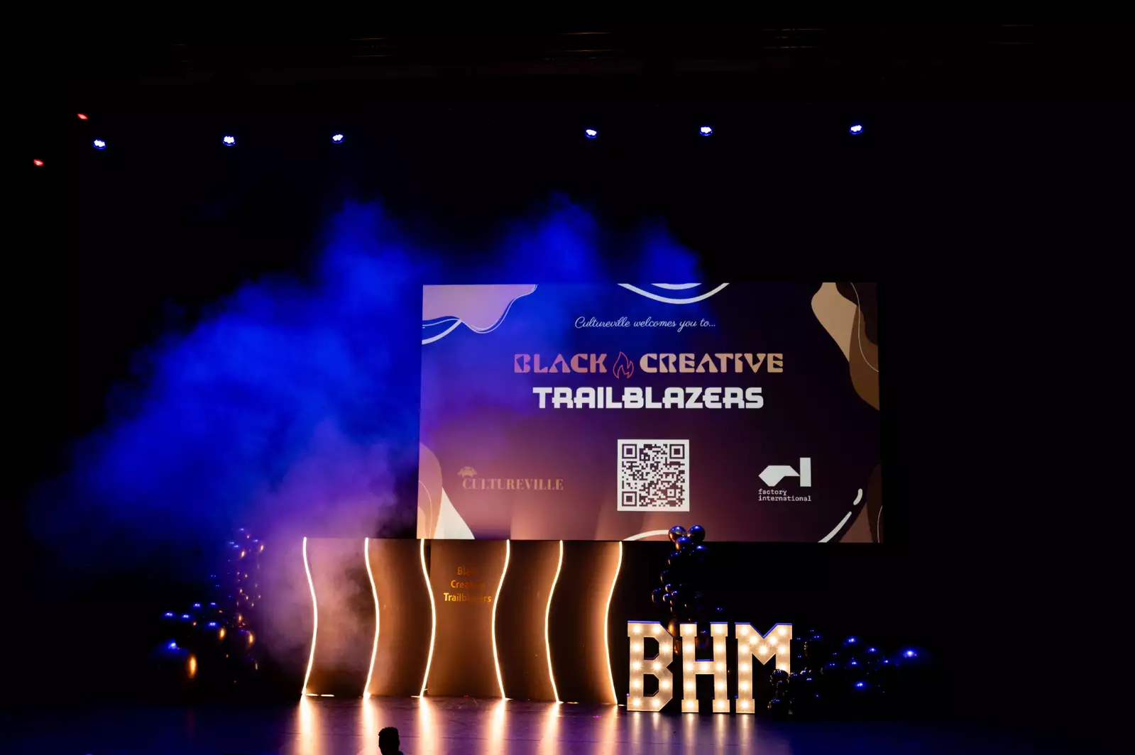 A stage is set with glowing decorations and a large screen displaying the title "Black Creative Trailblazers," hosted by Cultureville and Factory International. There is purple and blue lighting with smoke effects and balloon arrangements, and illuminated "BHM" letters celebrate Black History Month.