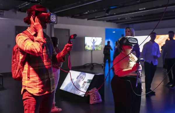 A man and a woman standing in a darkened art gallery, each wearing a Virtual Reality headset and holding a black controller. They are playing a game by the artist Rachel Maclean.