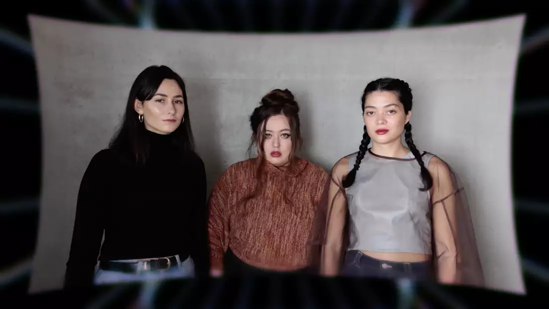 Three members of Keiken staring at the camera, stood in front of a large, grey piece of material.