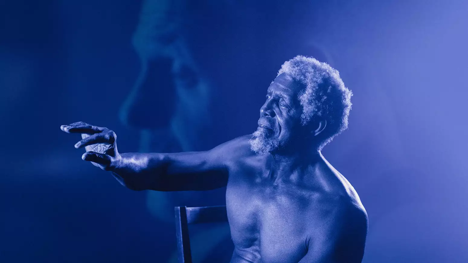 A shirtless elderly man with curly white hair and a beard sits on a chair, reaching out with an expressive hand. The image has a blue monochromatic tone with a soft-focus overlay of his face in the background.