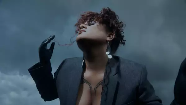 Lady Shaka wearing a grey blazer, black gloves, heart-shaped earrings, and sunglasses, looks upward against a cloudy sky. Their hand is raised near their face.