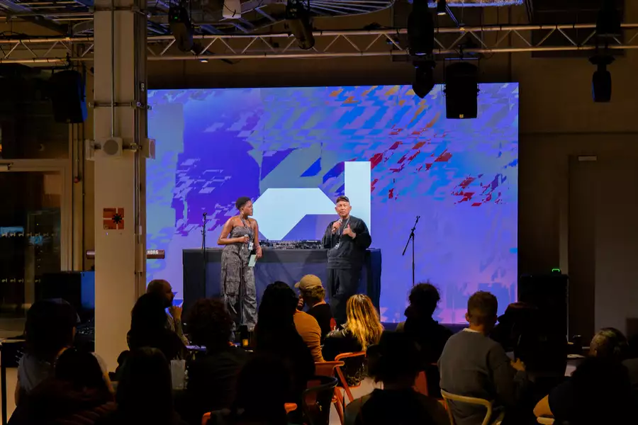 Two people stood on stage in front of DJ decks with the Factory International logo on a purple background on the screen behind them. There are a crowd of people sat on chairs watching them.