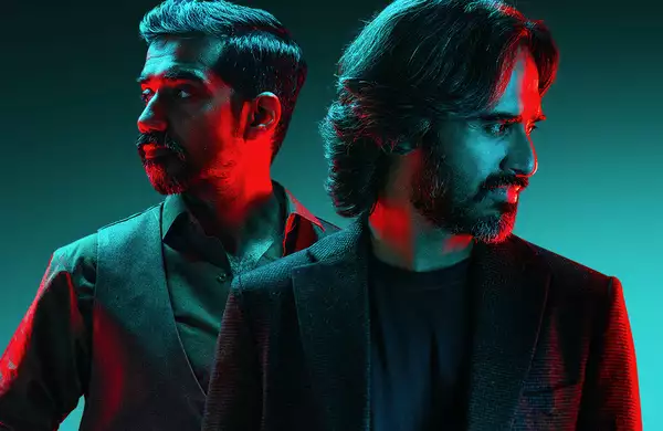 Two members of Kaavish standing back to back, looking in opposite directions, with dramatic red and blue lighting. The background is a gradient of teal.