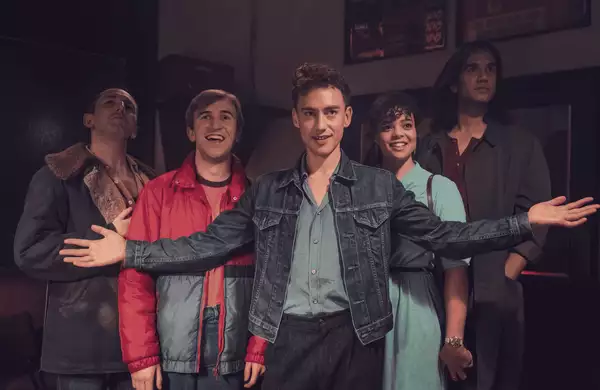 Cast image from It's a Sin showing five friends standing together indoors, smiling and looking upward, with the person in front wearing a denim jacket and holding their arms out wide.