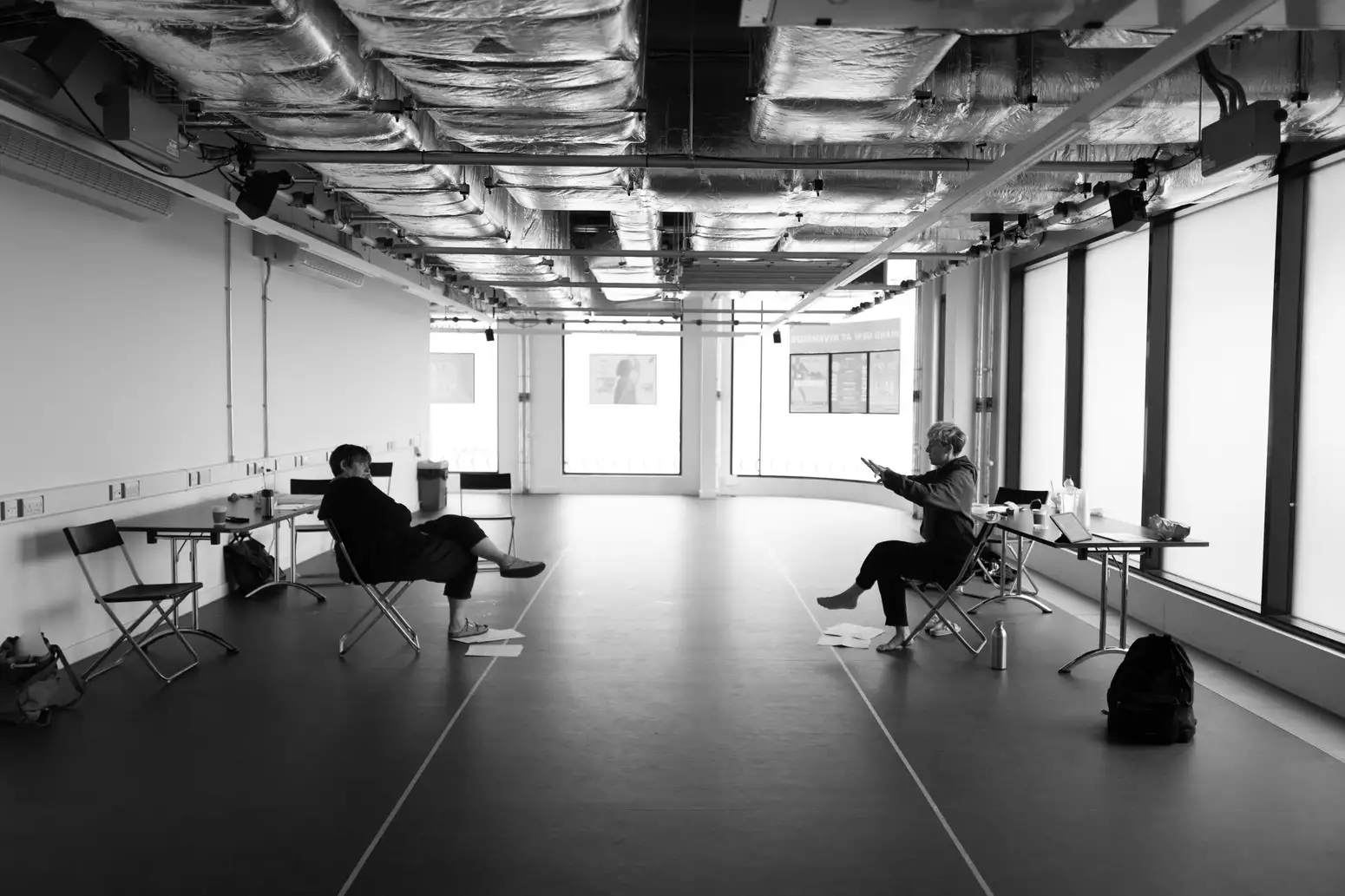 Black and white photograph of two people facing each other on chairs during THEY rehearsals