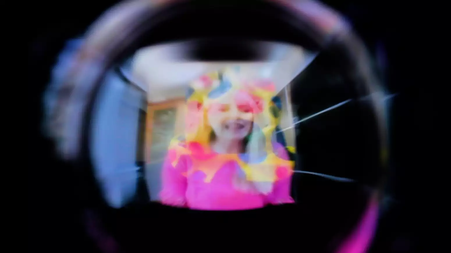 A blurred and light-distorted image down a dark camera lens of a smiling person with white skin wearing a bright pink top.