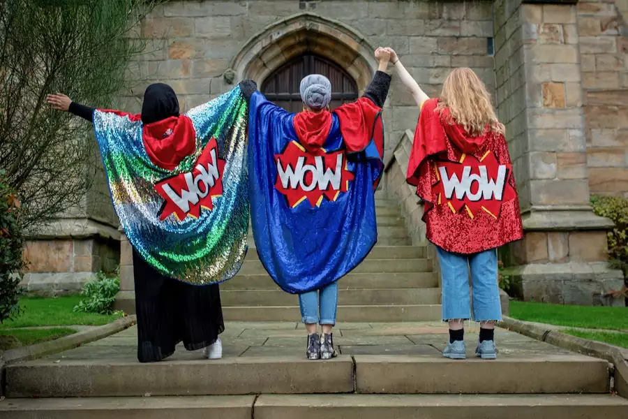 Three people with their backs turned and arms in the air, wearing glittery capes with the words 'WOW' on them