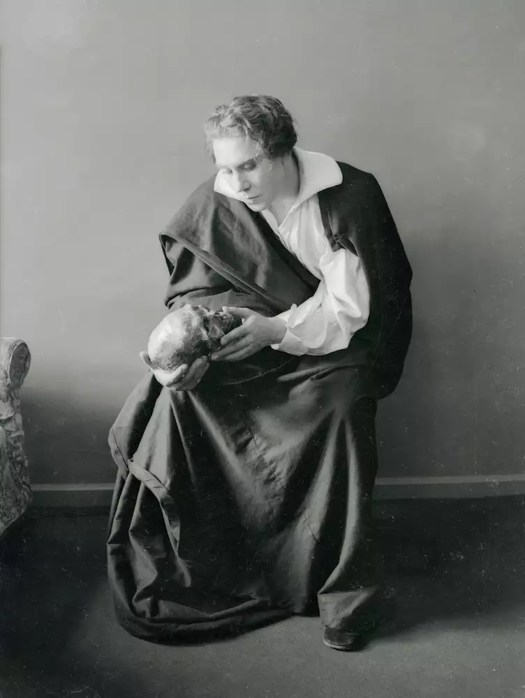 Black and white photograph of Donald Wolfit dressed as Hamlet in a gown. He is sat down and staring intently at a skull he is holding in his hands.