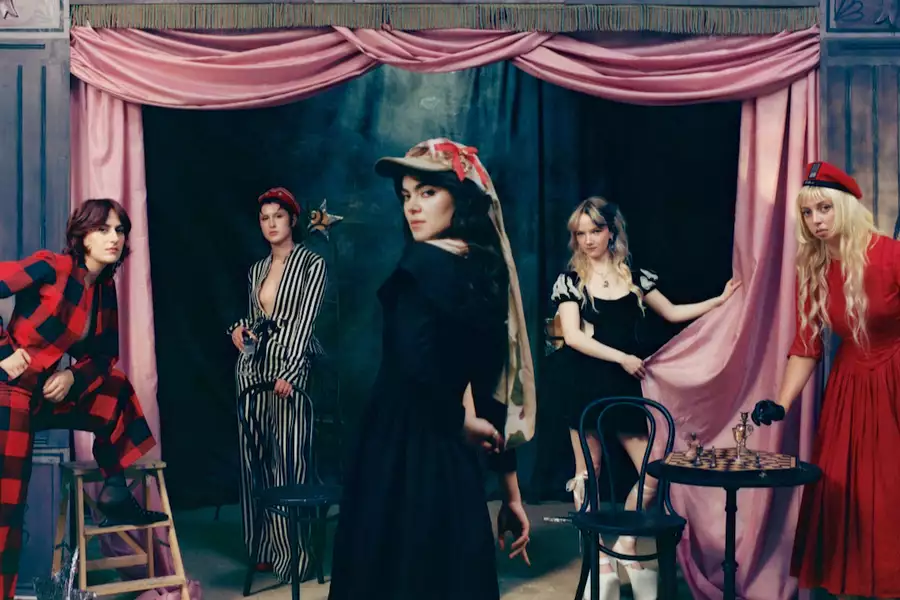 Five people from The Last Dinner Party in theatrical costumes pose on a staged set with pink curtains, including figures in black, red, and striped outfits around a small table with a chessboard.