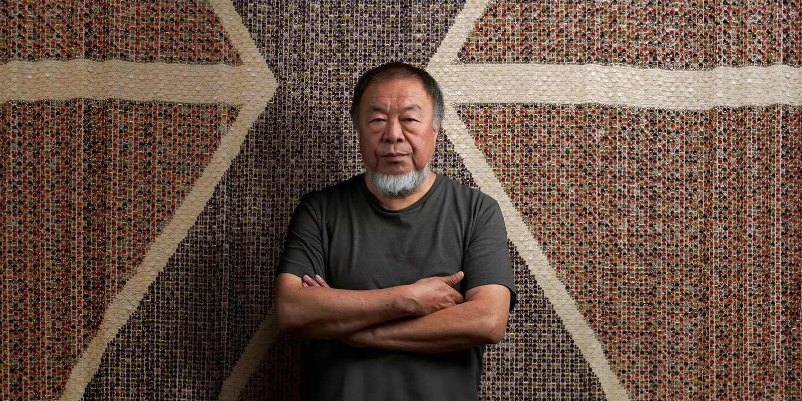 Ai Weiwei stood with arms folded wearing a black t-shirt, in front of a giant flag made of buttons