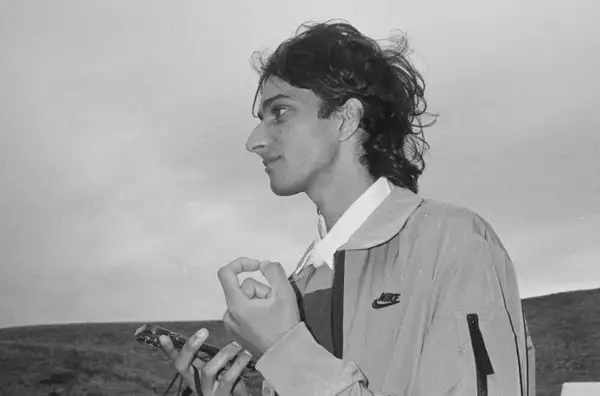 A black and white image of a young man in a rural landscape. He is looking past the camera to the left and wears a Nike jacket and holds a mobile phone in his right hand.