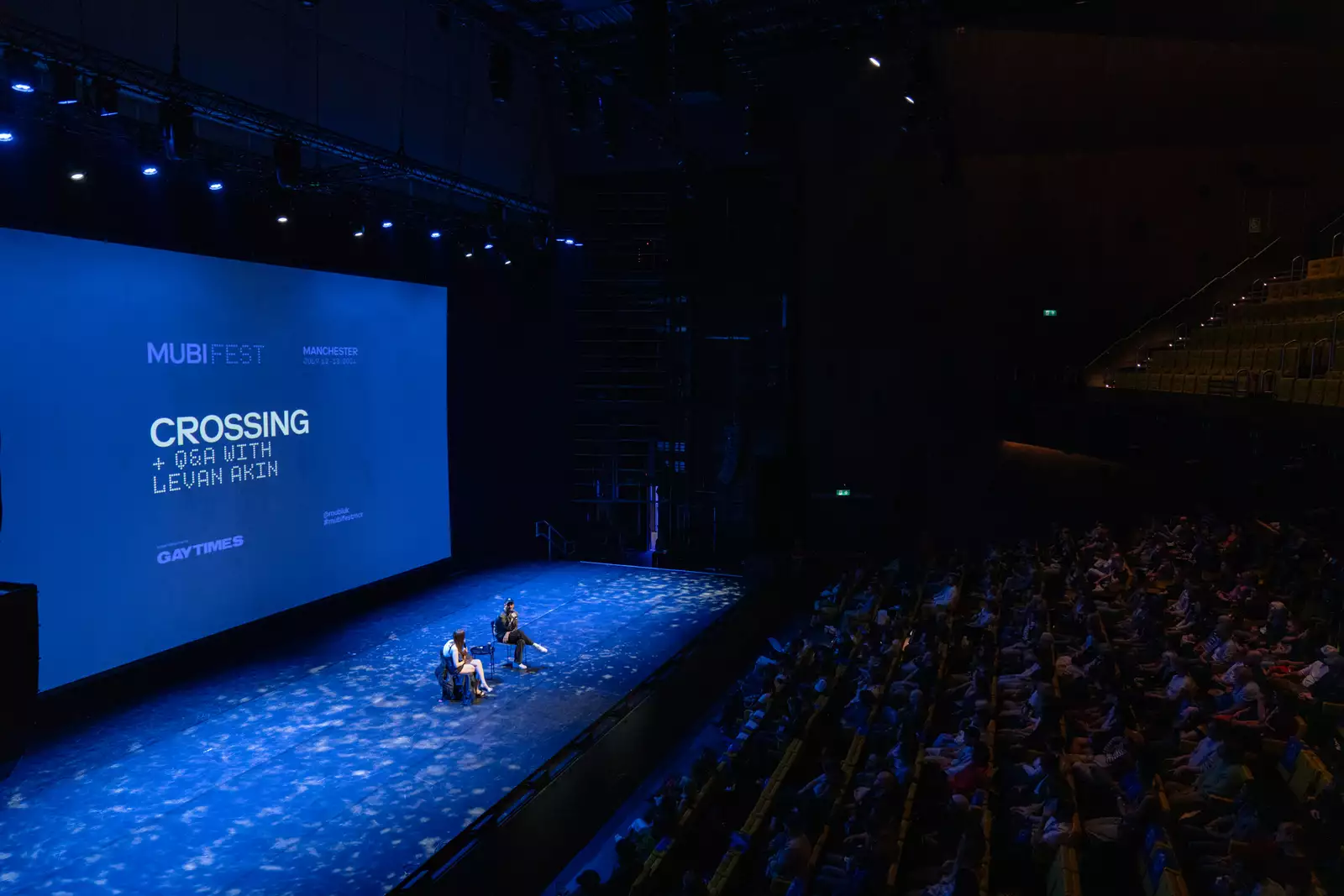 Film showing of 'Crossing' in the Hall. Two people are sat on stage for the Q&A in front of the big screen. The lighting is blue.