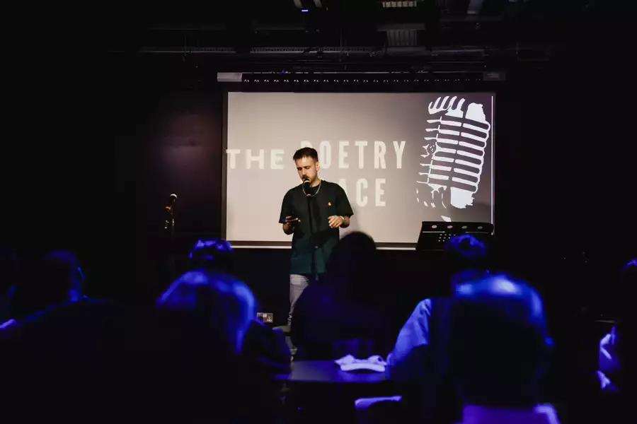 A poet performs spoken word poetry on stage in front of an audience, with a projection reading "The Poetry Place" behind him.