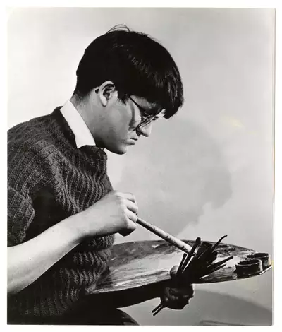 Black and white photograph of a young David Hockney holding an easel. He is wearing a jumper, shirt and glasses.