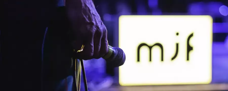 A close-up of a hand holding a microphone, with a glowing "mjf" sign in the background.